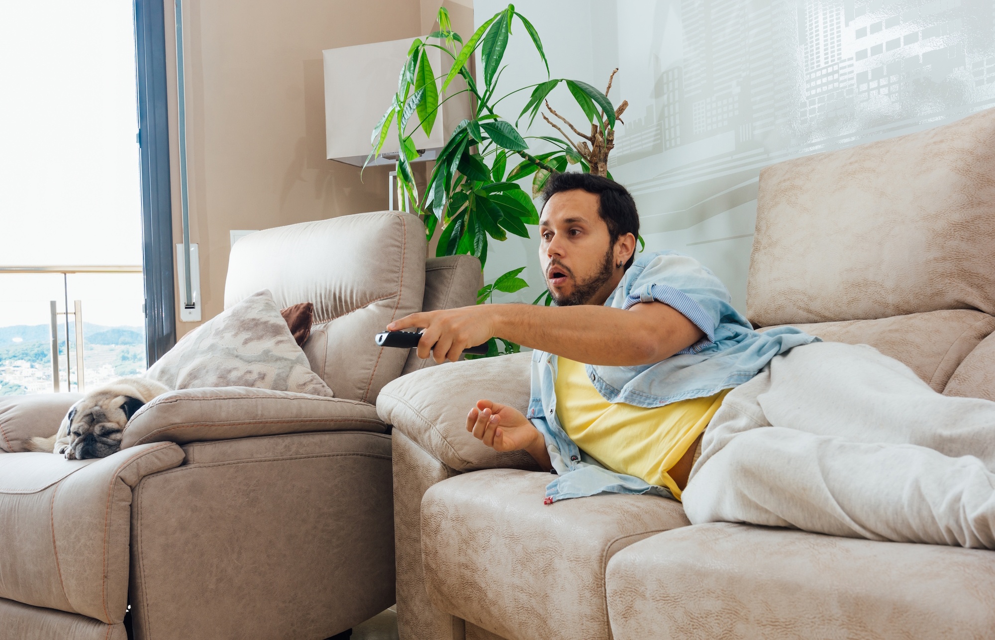 Homme allongé sur un canapé avec une télécommande - Les dangers du mode de vie sédentaire